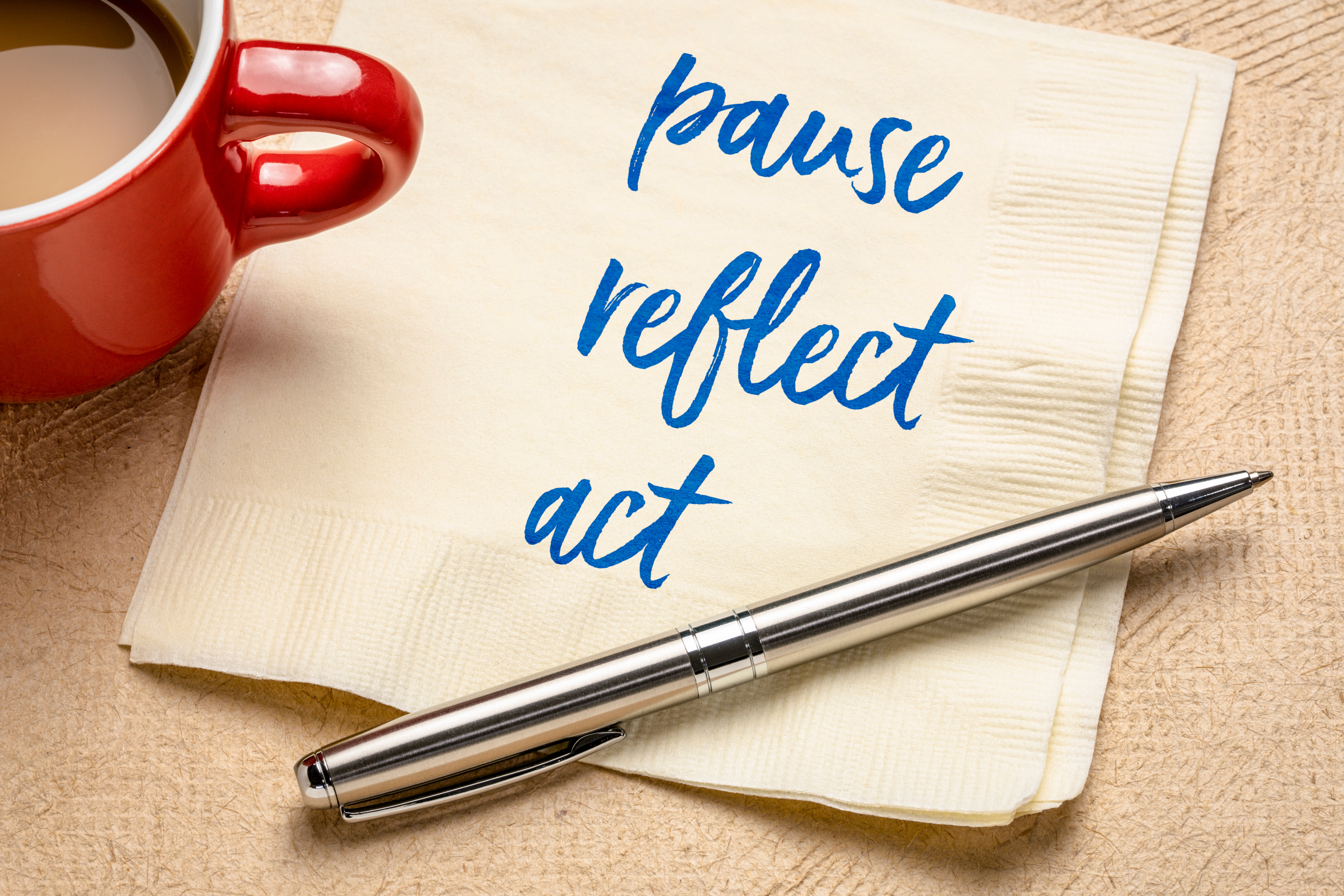 Coffee cup and a pen on a napkin with pause, reflect, act written on the napkin