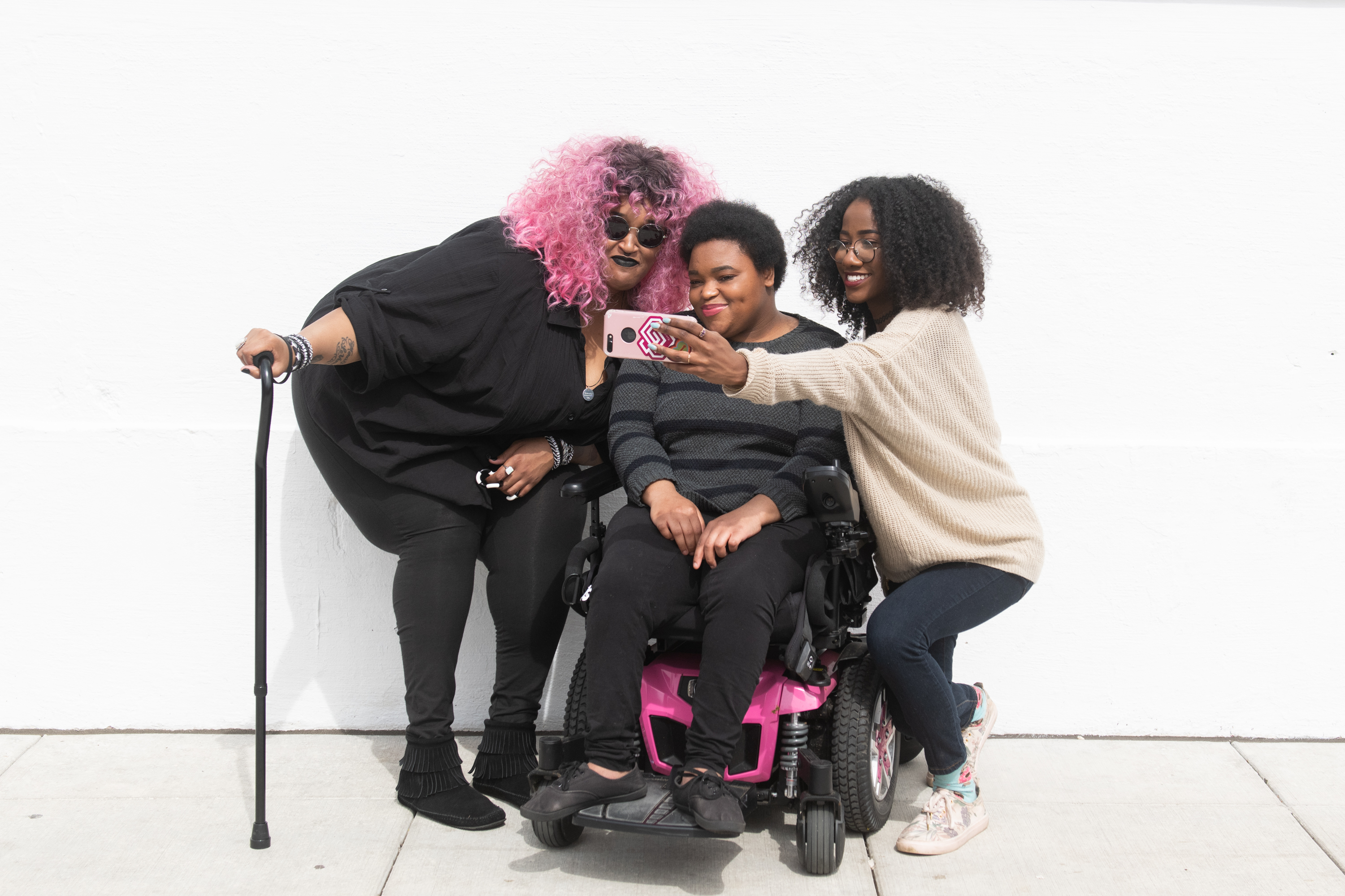 A zoomed out full body shot showing three Black and disabled friends (a non-binary person with a cane and tangle stim toy, a non-binary person sitting in a power wheelchair, and an invisibly disabled femme) smiling and taking a cell phone selfie together. All are outdoors in front of a white wall. Attribution: Disabled and Here Collection.