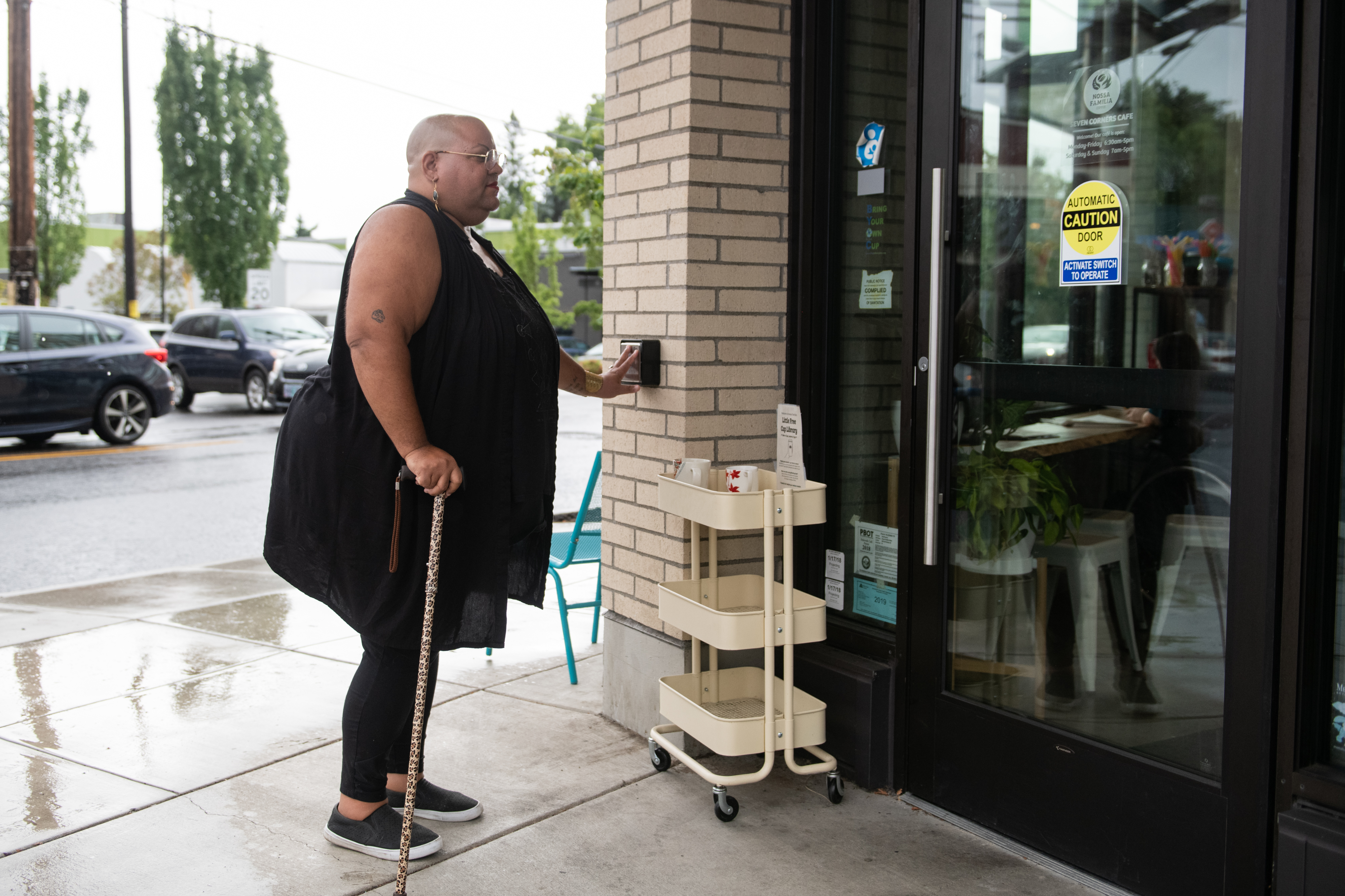 A Black non-binary person with a leopard print cane presses a wall-mounted push button to open the cafe door. They are dressed in all black and have a shaved head, glasses, and a red lip on. Attribution: Disabled and Here collection.