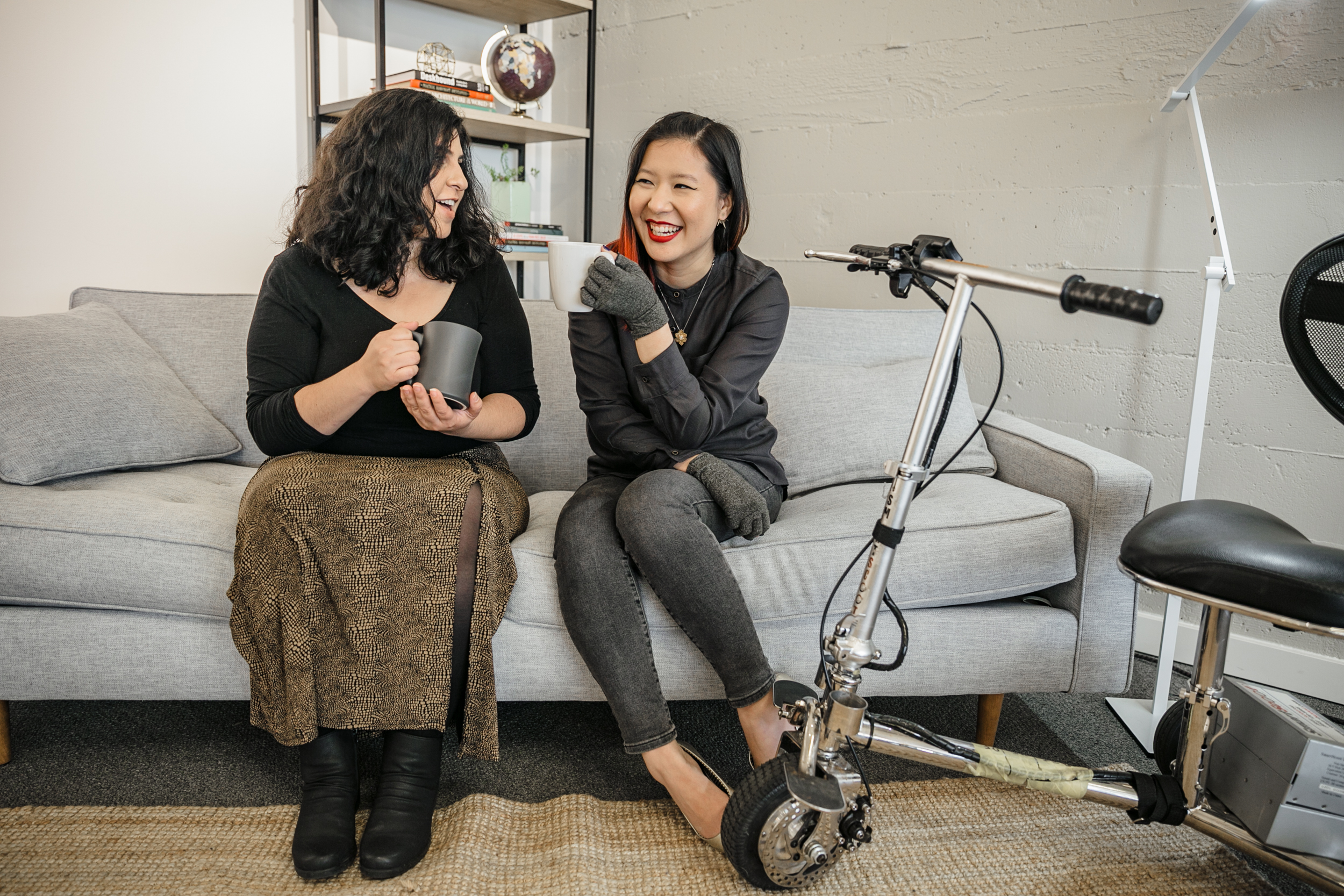 A Latinx woman with an invisible disability and an Asian disabled genderfluid person chat and sit on a couch, both holding coffee mugs. An electric lightweight mobility scooter rests on the side. Attribution: Disabled and Here collection.