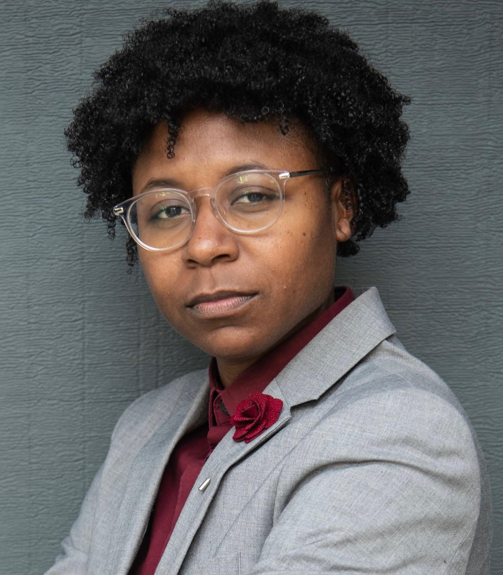 B, a Black non-binary human, is pictured in front of a grey backdrop wearing clear glasses frames and a grey suit jacket over a dark red shirt with a red rose on the lapel. Their curly hair is worn free. 