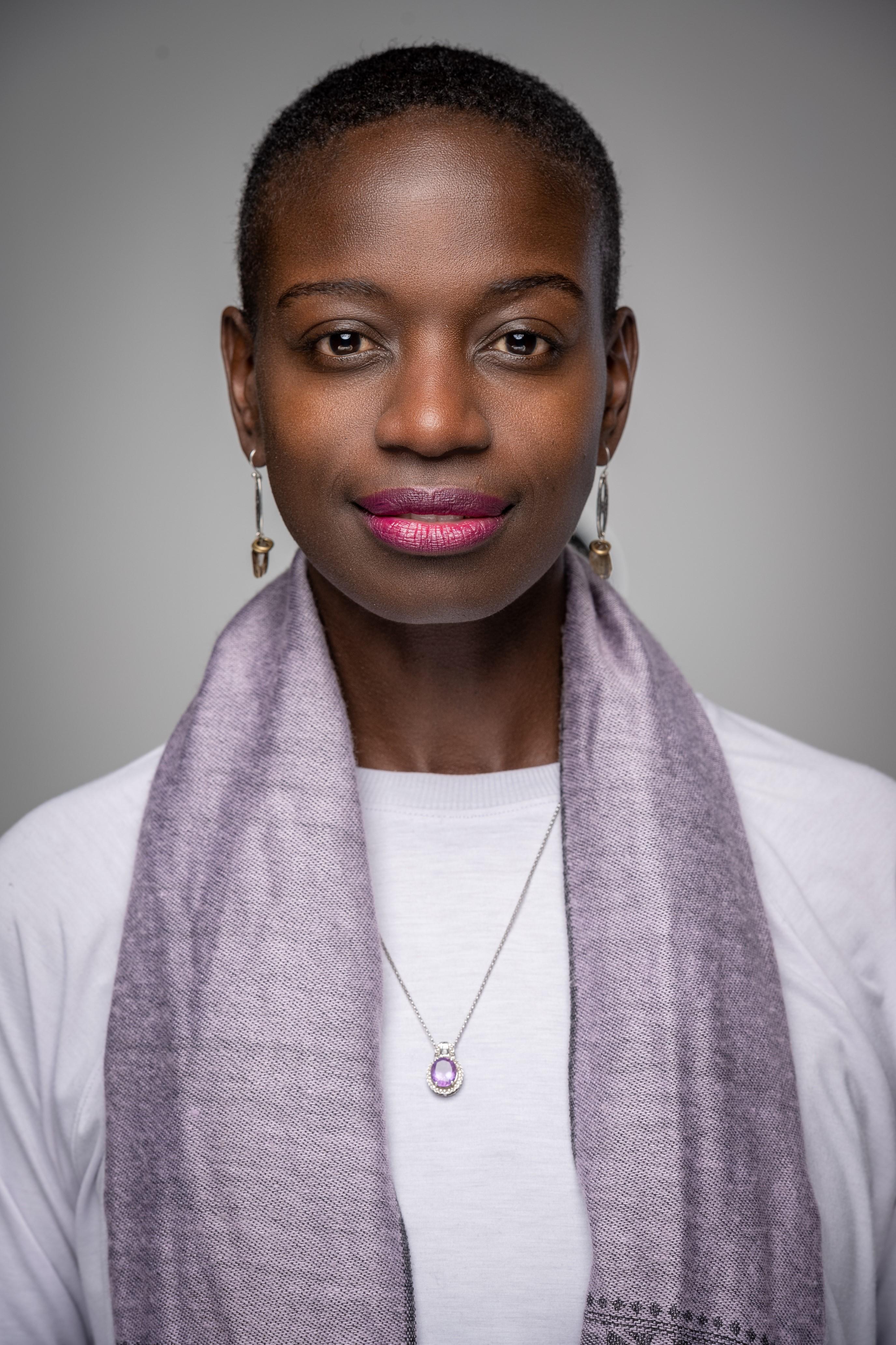 This is a professional photo of a Black businesswoman, with short hair. She is wearing a lavender shirt and scarf. The background is slate grey. The photo is taken from the waist and above.   