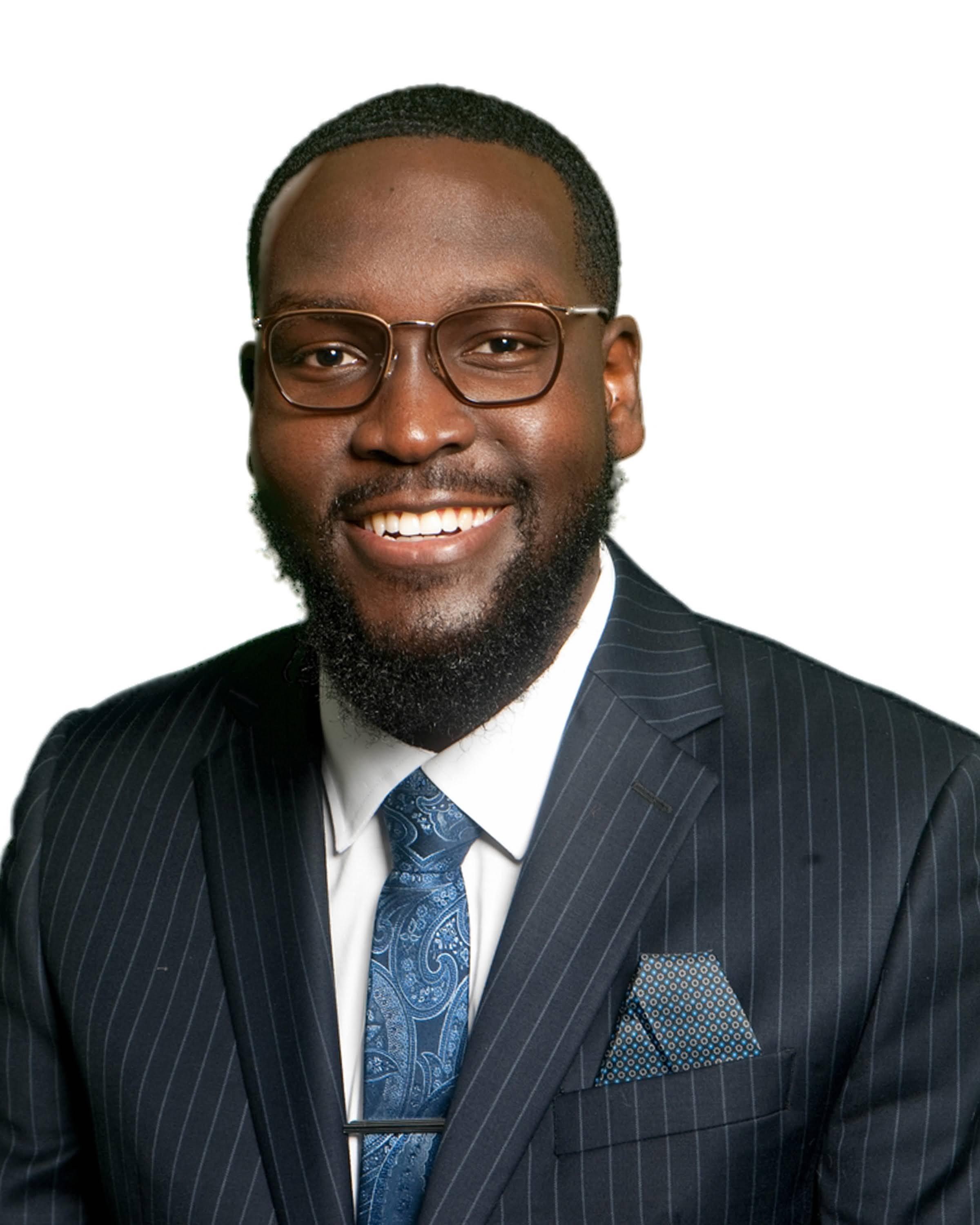 Jean, a Black man, is wearing white shirt, blue tie and a navy blue blazer with light color stripes. He wears glasses, has short hair and beard.   