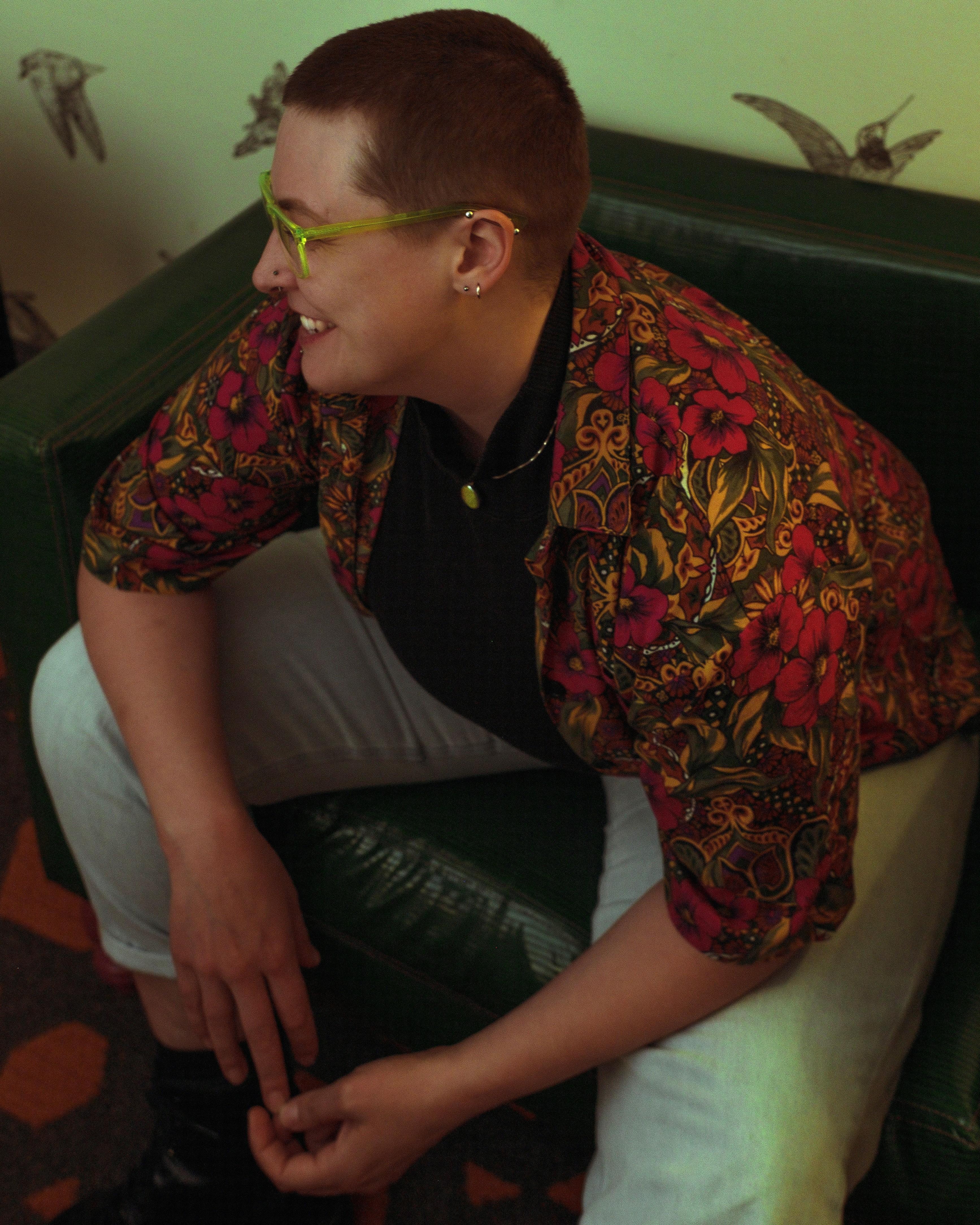 Rook, a white non-binary trans person, looks away from the camera smiling. They’re wearing jeans, a black turtleneck, and a colorful button up t-shirt. They have a multitude of facial piercings and neon green jewelry and glasses. 