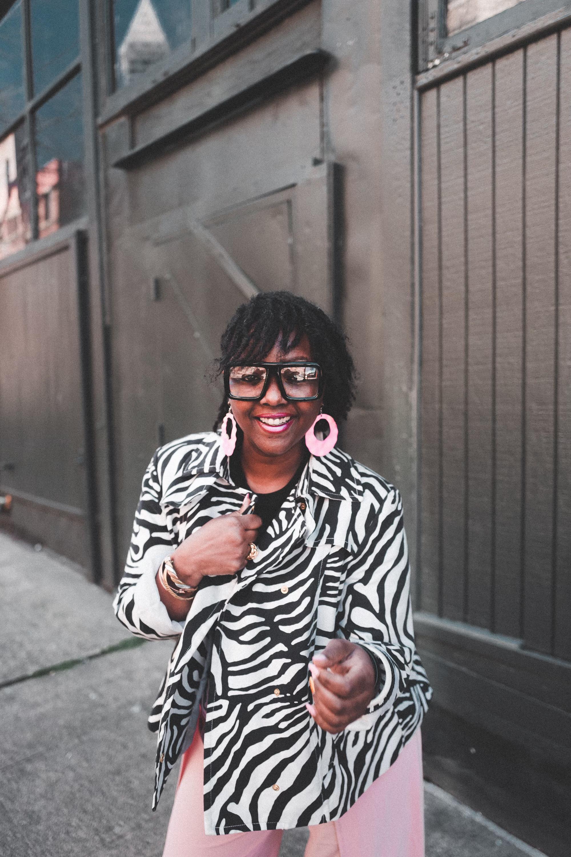 TraeAnna, a Black woman, stands in front of an industrial background wearing a black and white zebra print coat with pint pants. She is smiling and her locs hang loose around her face. 