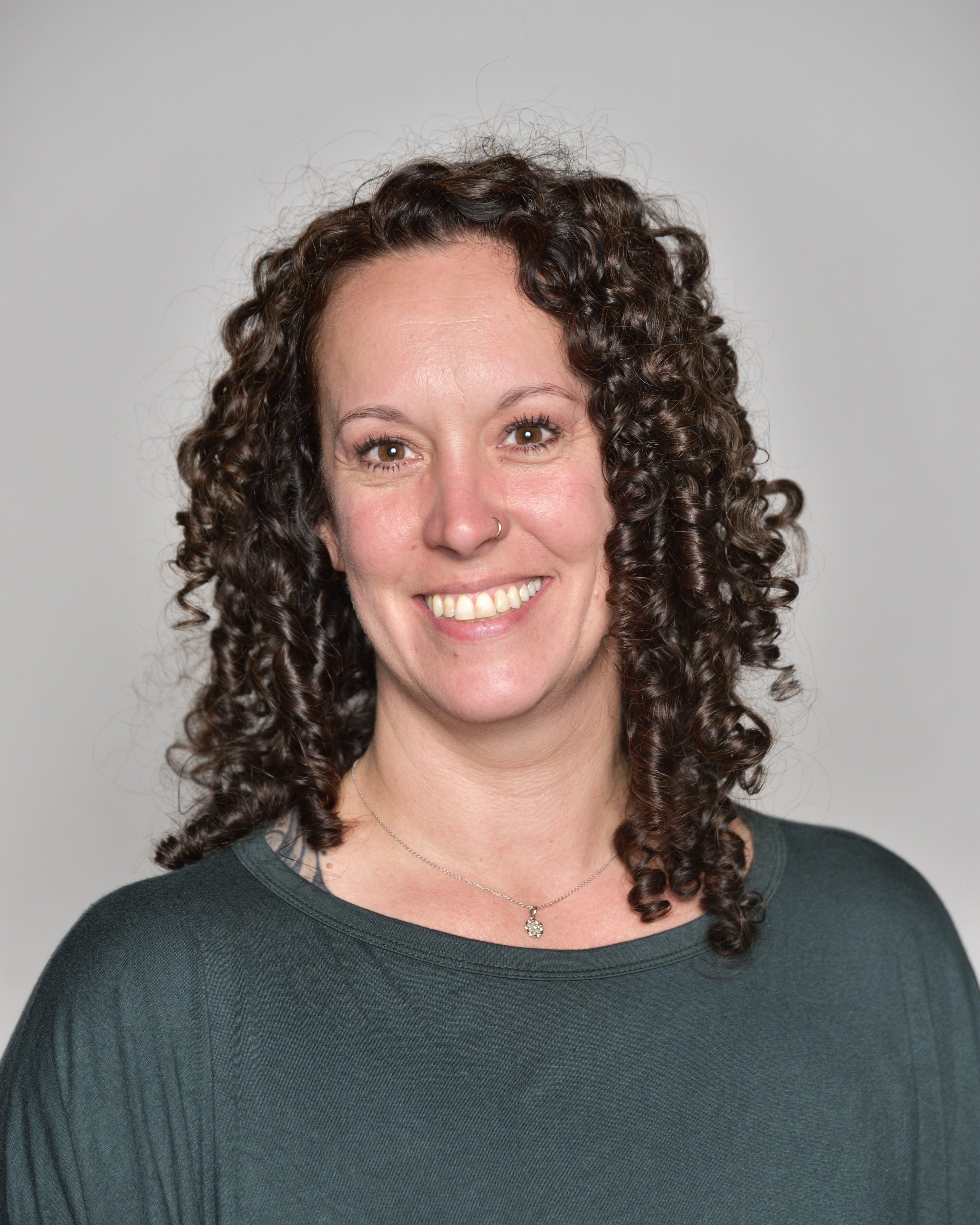 A woman with curly brown hair and wearing a green top smiling at the camera in front of a off-white plain background. 