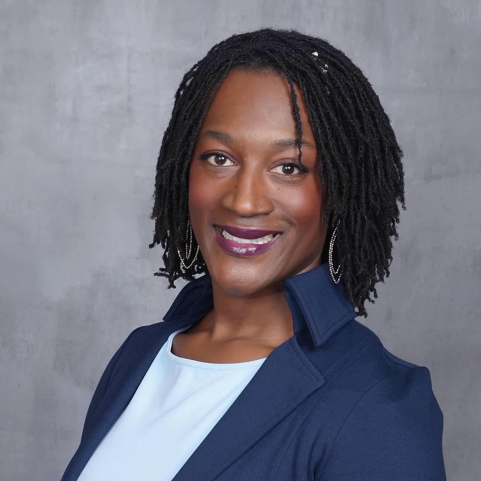 Megan, a Black woman, smiles at the camera, her shoulder length sisterlocks are worn loose around her face. She wears an orange button up shirt underneath a black blazer. 