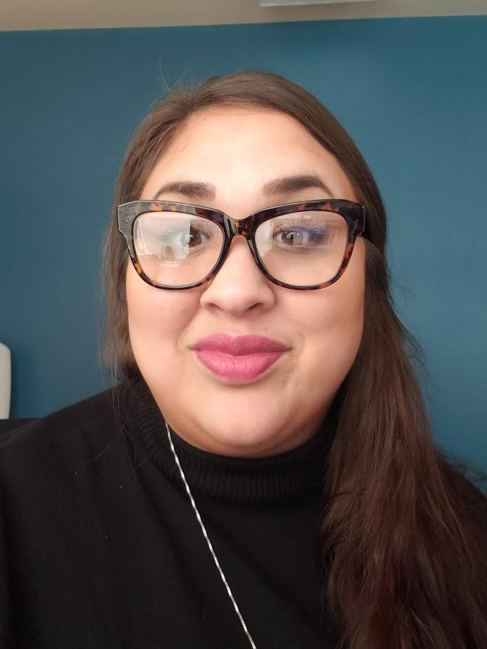 Woman with brown hair stands in front of blue background wearing black turtleneck and tortoiseshell glasses. 