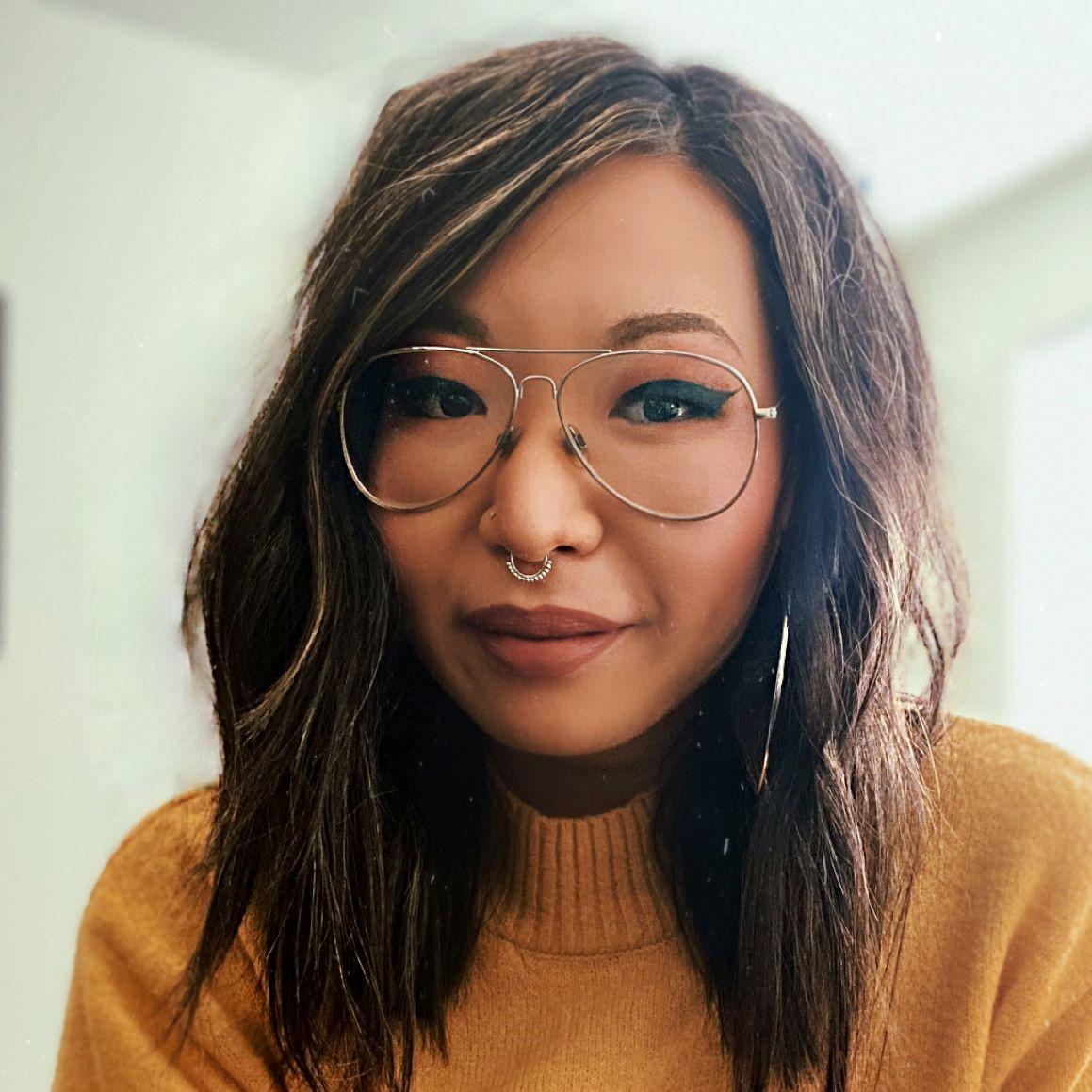 A woman from the shoulders up with long, wavy dark brown hair parted on the side with golden highlights wearing gold rimmed glasses, black winged eyeliner, and gold jewelry in her nose with full lips in a relaxed smile wearing a deep yellow sweater against a soft white background.