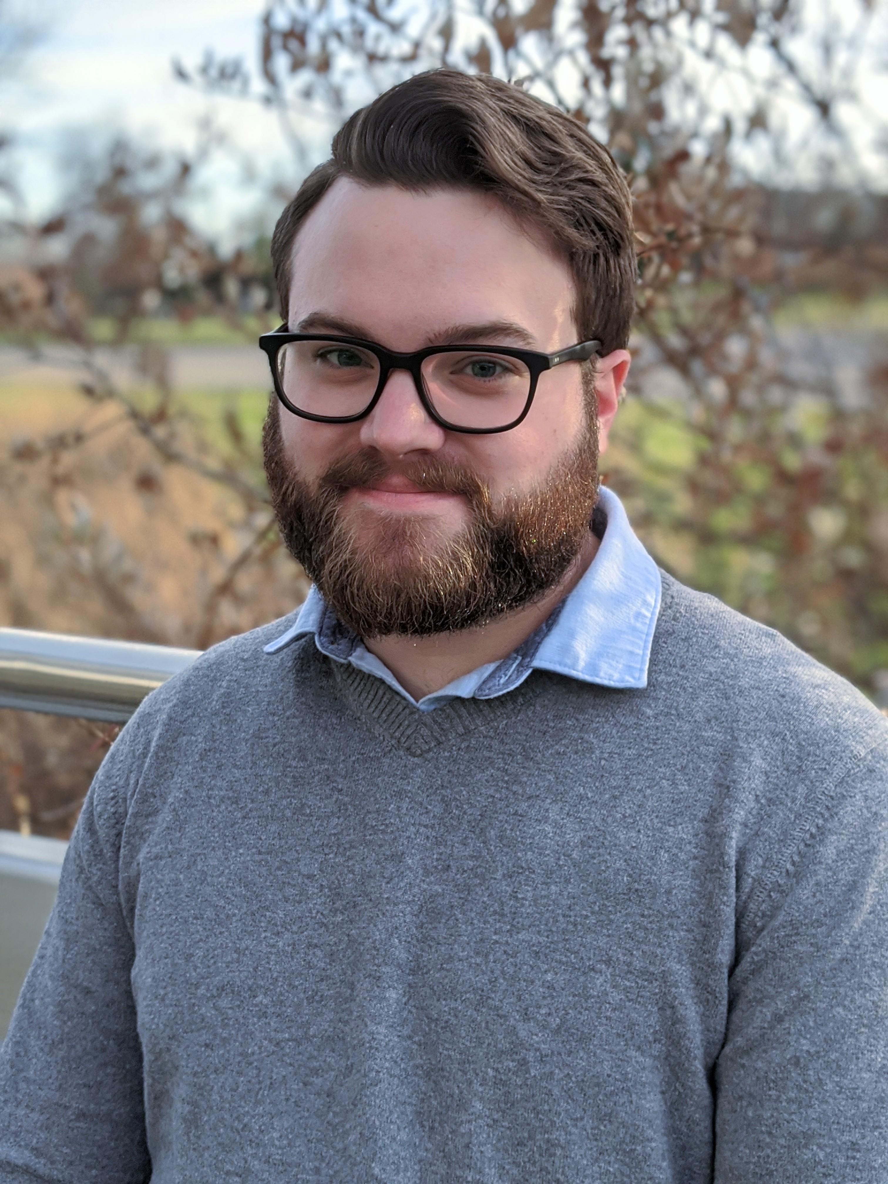 A man from the chest up wearing dark rimmed glasses with a wave of short brown hair, a full, auburn mustache and beard, and a relaxed smile wearing a grey V-neck sweater over a light blue collared shirt in front of a softly blurred background of natural brush, grass, and sky.