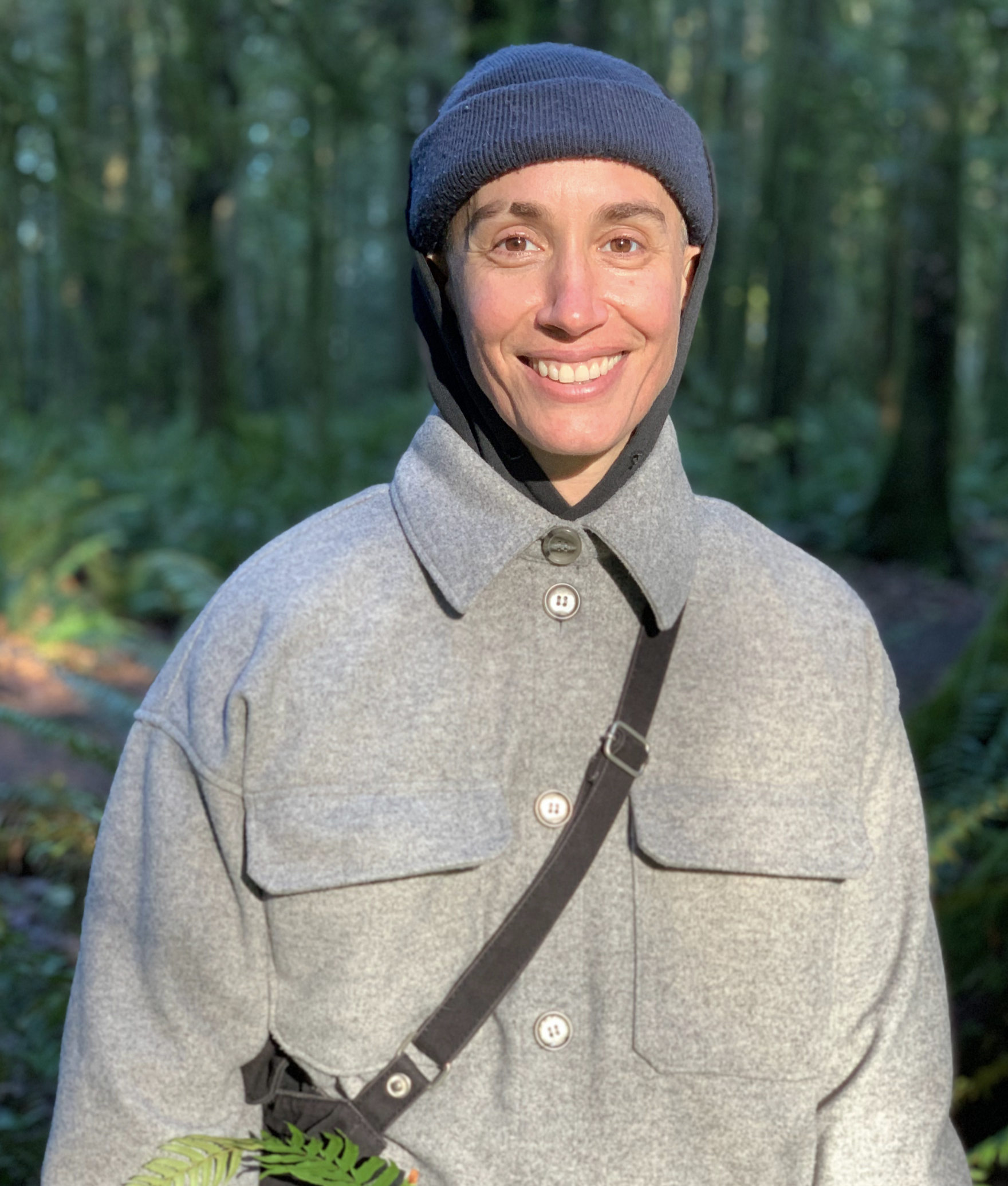Person with gray button up long sleeve coat iwith a brown sling bag over their shoulders. They are wearing a blue beanie and a wooded area is in the background.