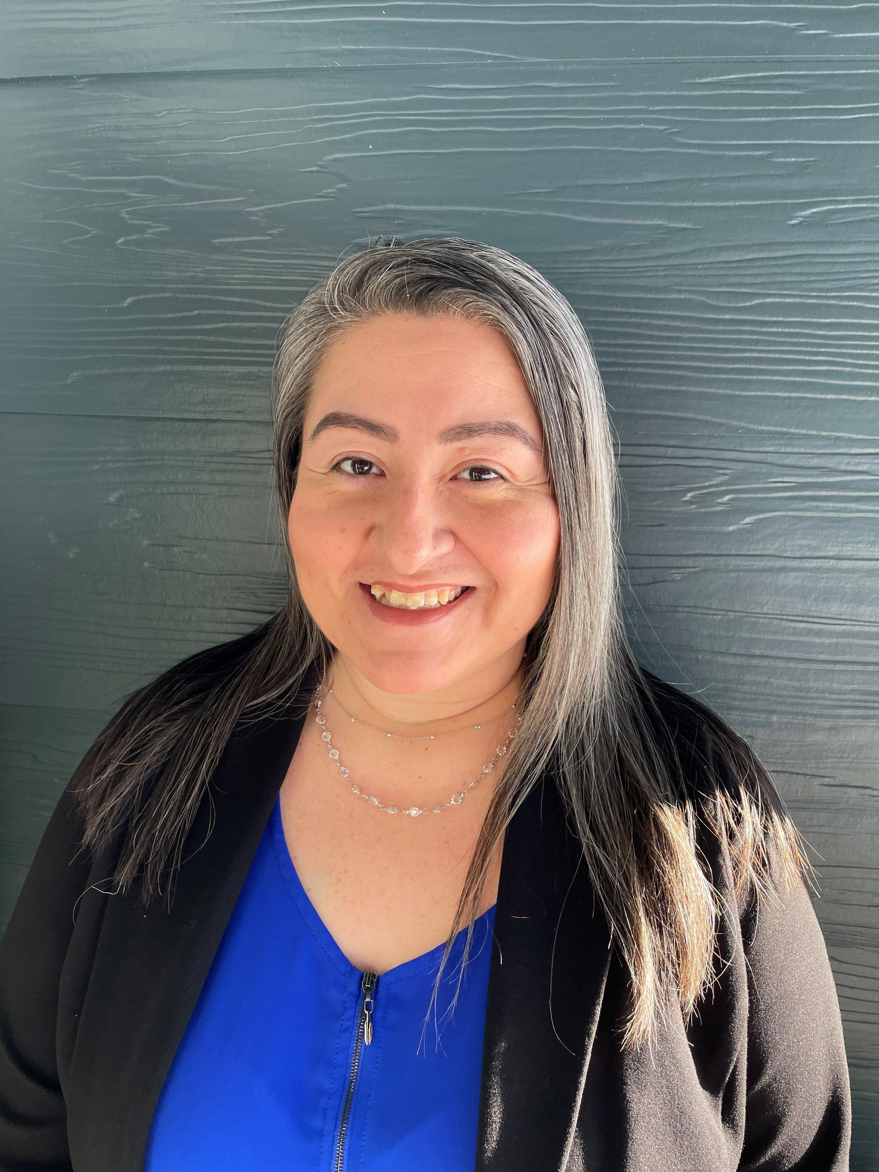 Yeni, a white presenting Latine woman, is pictured in front of a teal wall wearing a black cardigan over a blue shirt. Her long grey hair is worn loose about her face and she is smiling.  