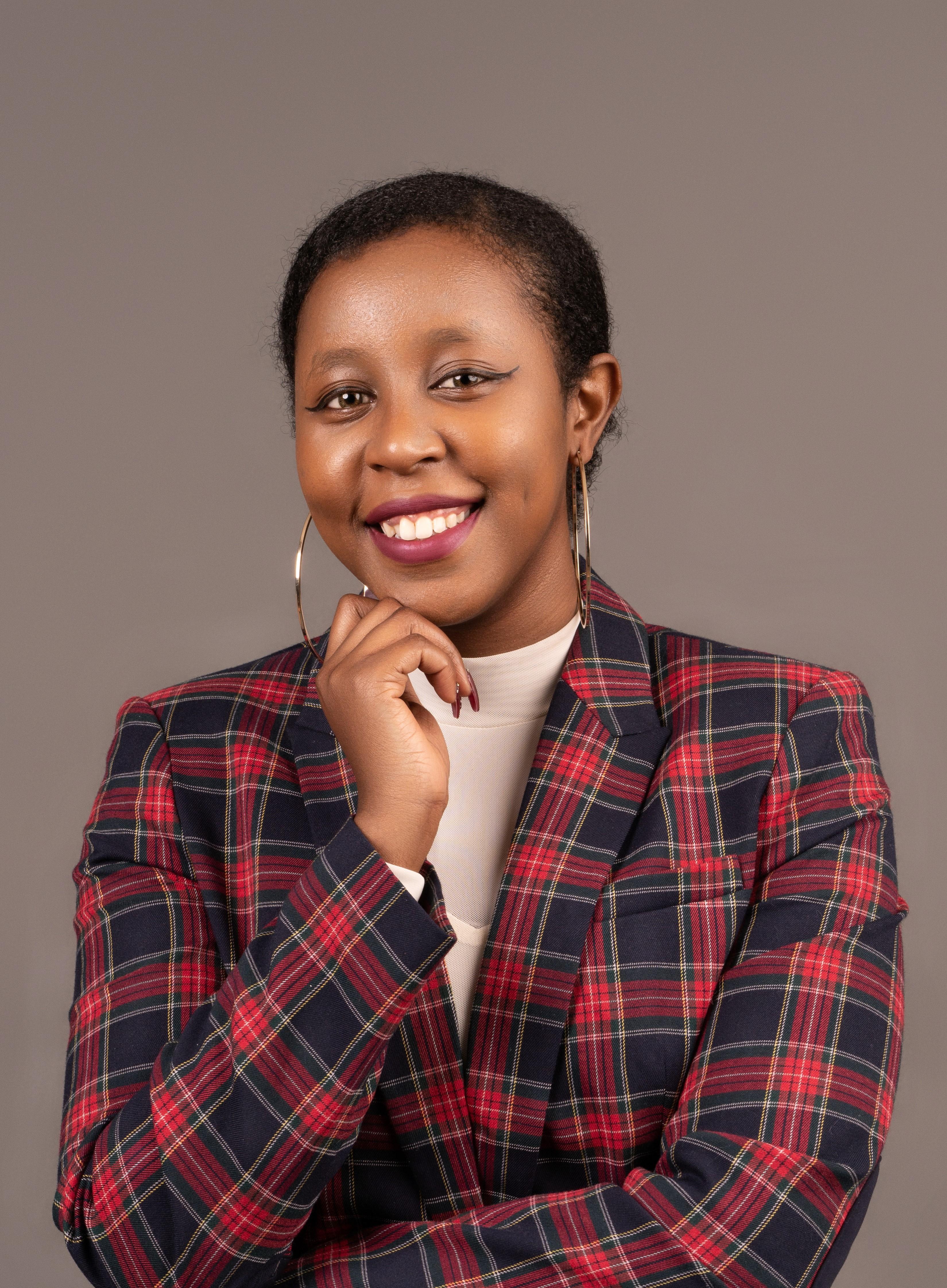 A woman with brown skin, black hair, and winged eyeliner smiles against a taupe background. She wears a plaid blazer, cream shirt, blue jeans, and hoop earrings.