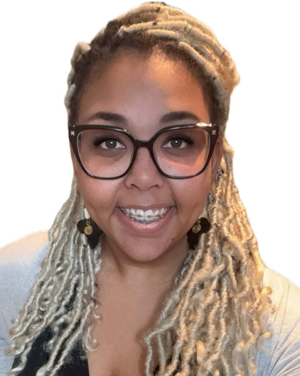 A woman with light brown skin, deep brown eyes, and braces smiles against a white background. She wears black glasses, Africa-shaped earrings, and has long blonde locs.