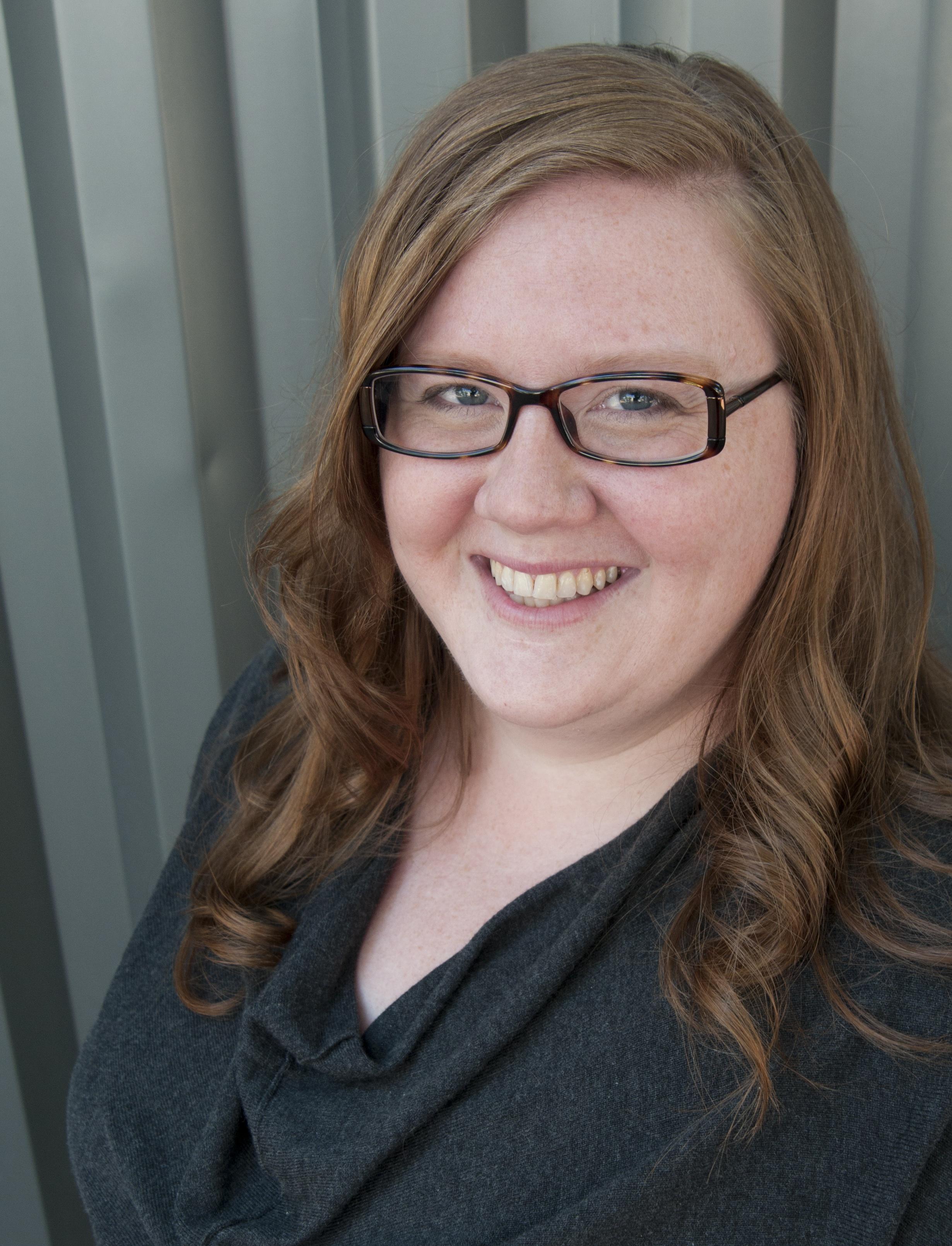 A woman with light, freckled skin, blue eyes, and auburn curls smiles against a greenish-grey background. She wears a dark grey cowl-neck sweater and brown glasses.