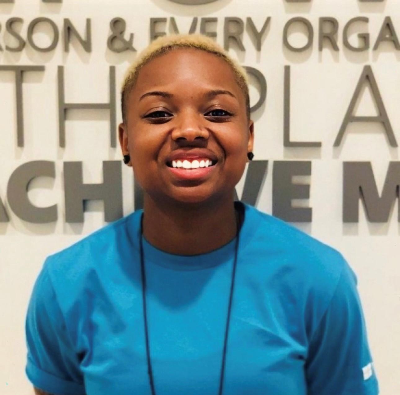 A woman from the chest up with her shoulders leading forward and her chin raised slightly stands before a cream background with taupe lettering. She has deep brown skin and dark eyes with full lips and a bright smile. She has short natural hair with a blonde crown and black stud earrings, and she is wearing an aqua blue t-shirt.