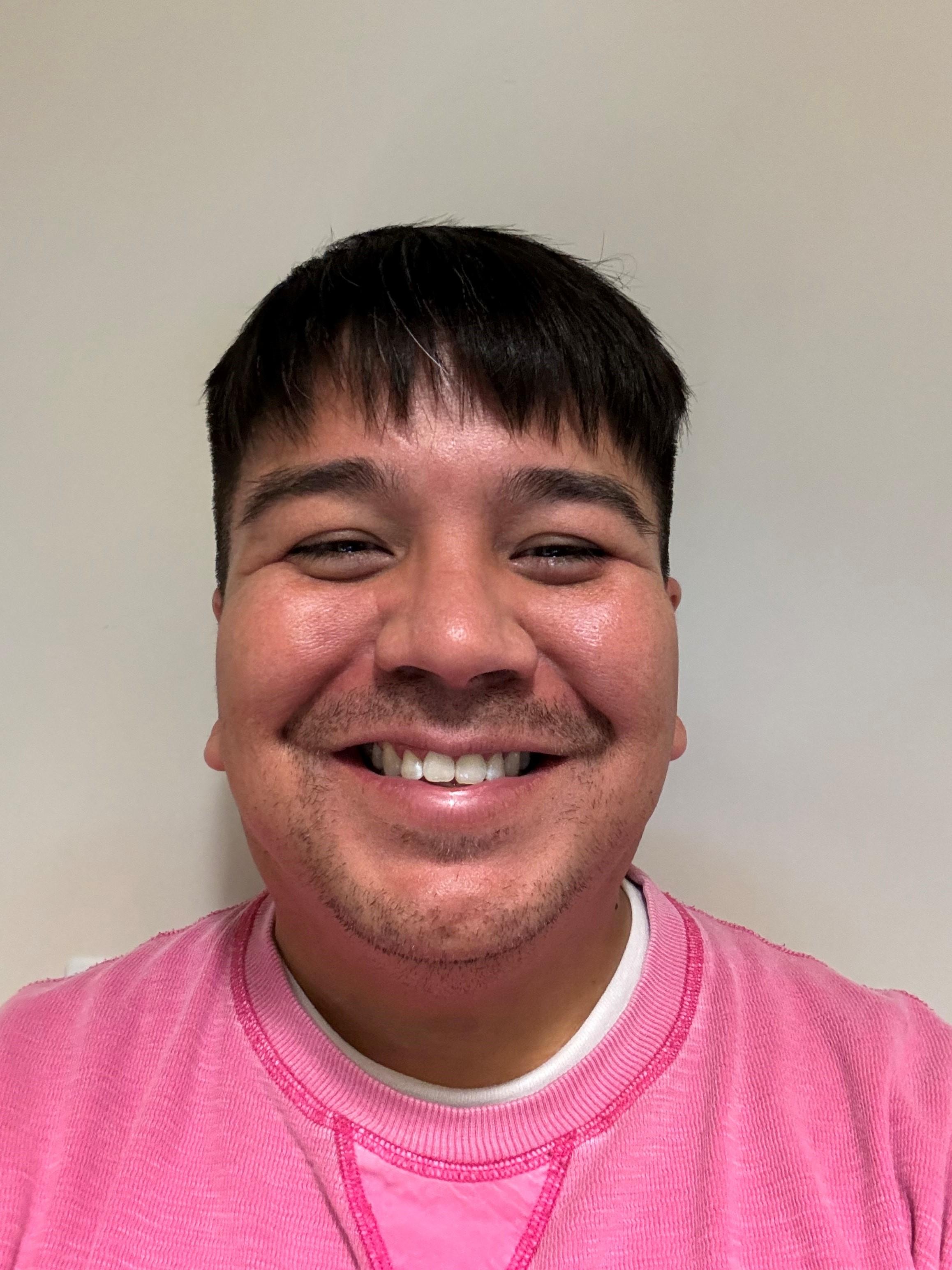 A man with light brown skin, short black hair, goatee shadow, smiling with rosy cheeks, in a bright pink shirt over a white t-shirt, against a taupe background.