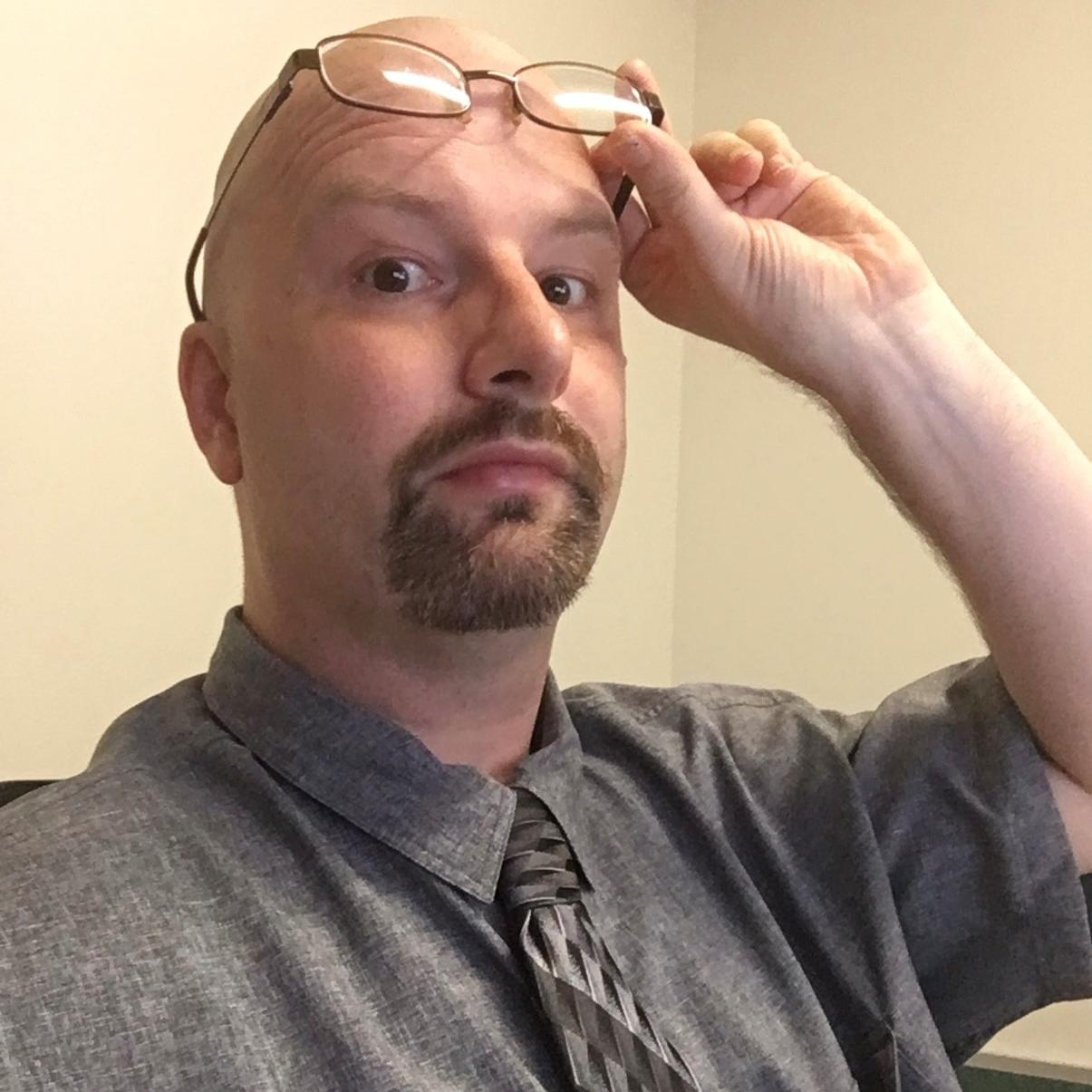 Photo of a man with a shaved head sitting in a chair and lifting his glasses to his forehead. He is wearing a gray dress shirt and tie and is in front of a tan background.