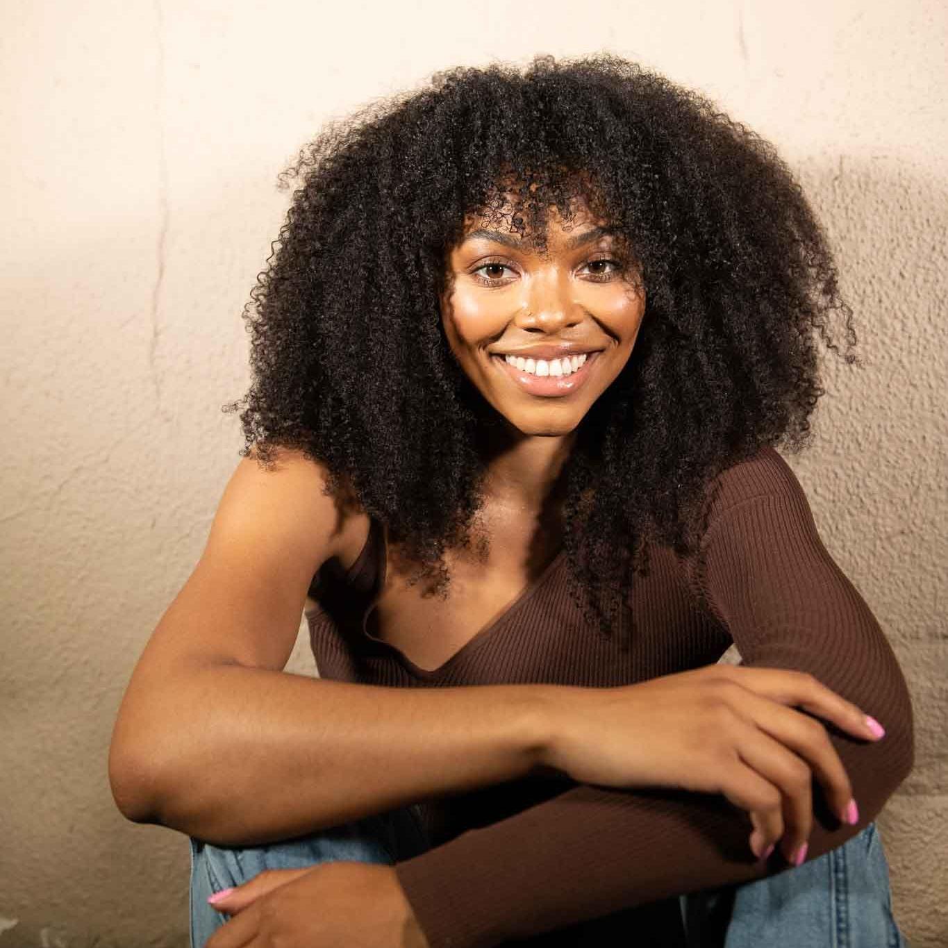 Woman with long brown hair wearing a brown top that exposes the shoulder and blue jeans sitting in front of a beige textured wall. She is looking at and smiling at the camera.