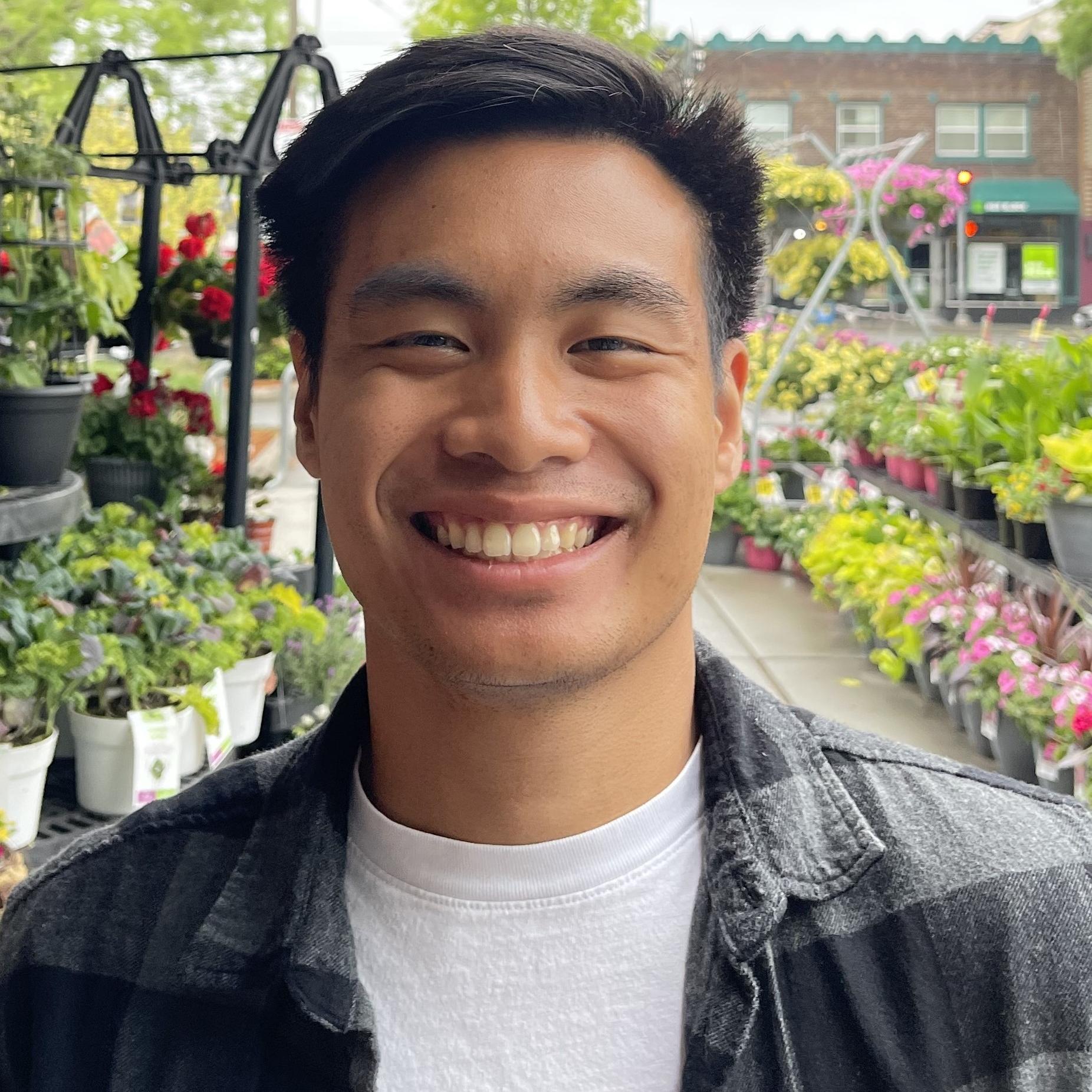 Close up shot of a person with tan skin and black hair smiling at the camera. They are wearing a white t-shirt with a flannel over it, and they are standing in a display of flowers.