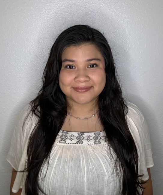 Image of woman with long black hair in a white top smiling at the camera. She is standing in front of a white textured wall. 