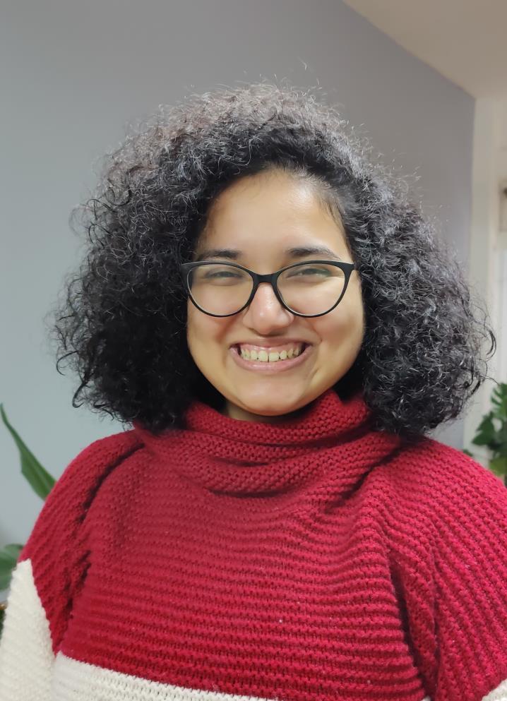 Person with tan skin wearing black-rimmed glasses. They are smiling at the camera. They have curly black hair to their shoulders and they are wearing a baggy red sweater with a white stripe near the stomach. 