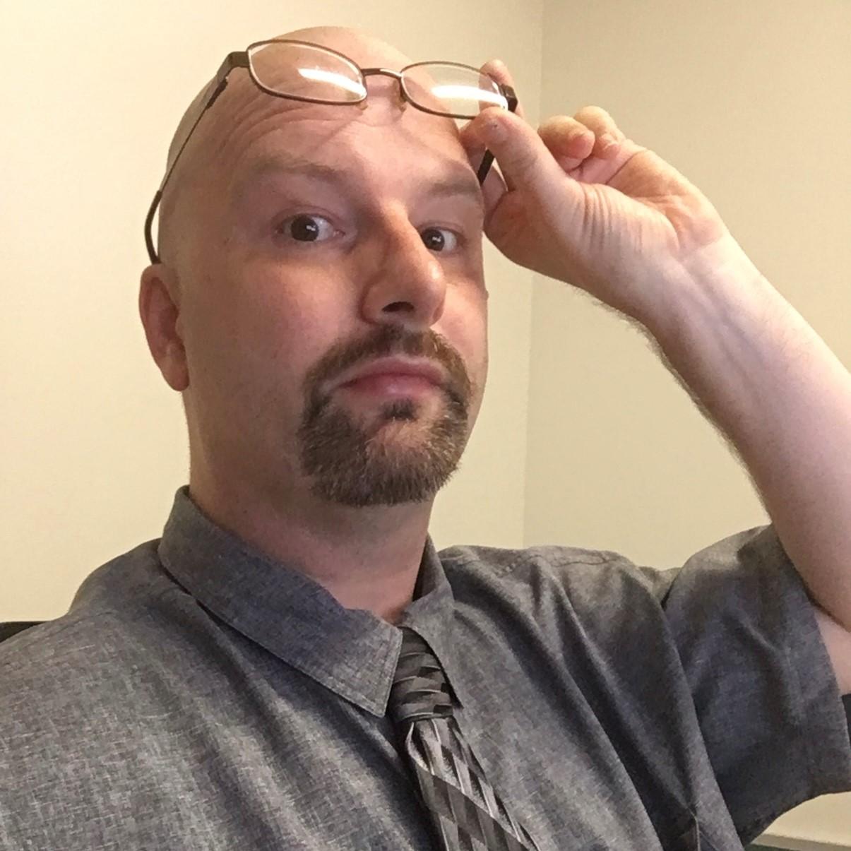 Photo of a man with a shaved head sitting in a chair and lifting his glasses to his forehead. He is wearing a gray dress shirt and tie and is in front of a tan background.