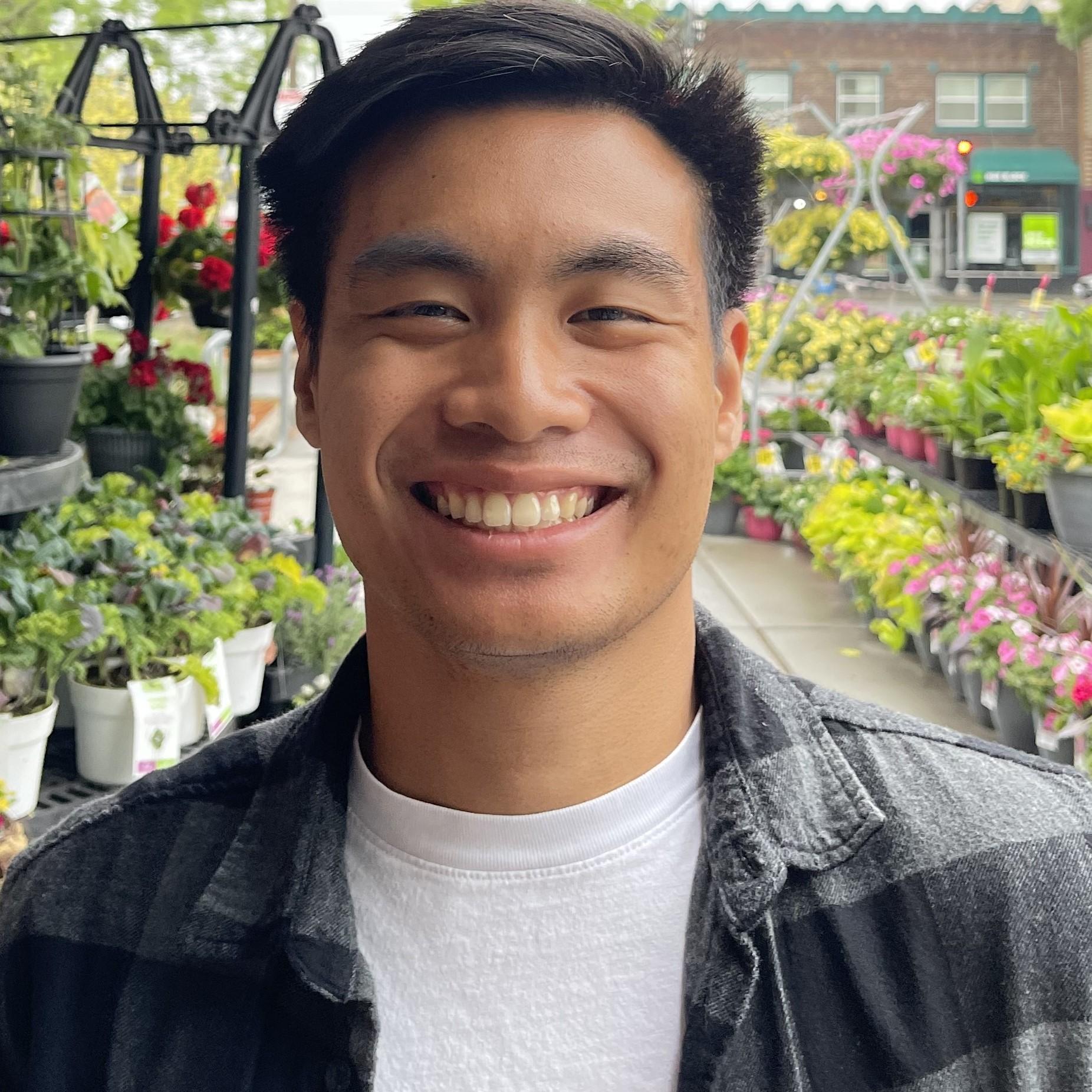 Close up shot of a person with tan skin and black hair smiling at the camera. They are wearing a white t-shirt with a flannel over it, and they are standing in a display of flowers.