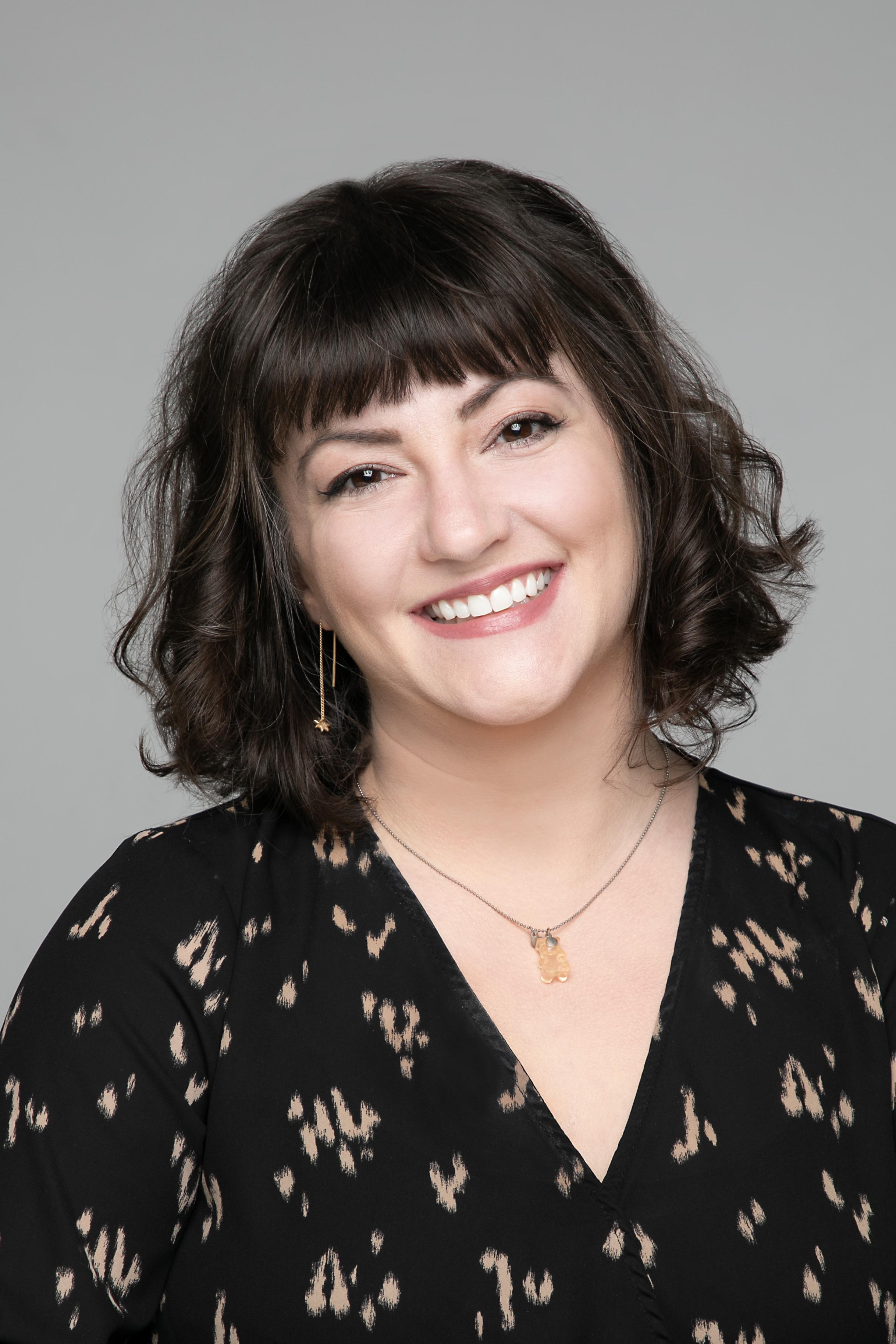 Photo of a smiling person with shoulder length black hair and dark eyes. they are wearing a black patterned top and a gold chain necklace.