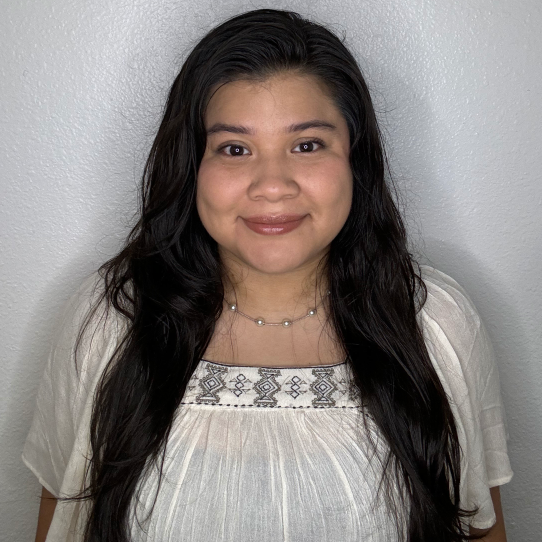 Image of woman with long black hair in a white top smiling at the camera. She is standing in front of a white textured wall. 