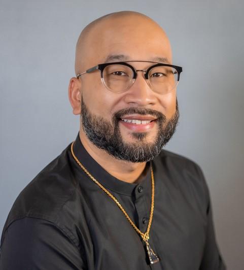 Photo of a bearded man with a shaved head smiling at the camera. They are wearing a black button up top and a narrow gold chain necklace with a triangle-shaped charm hanging from it.