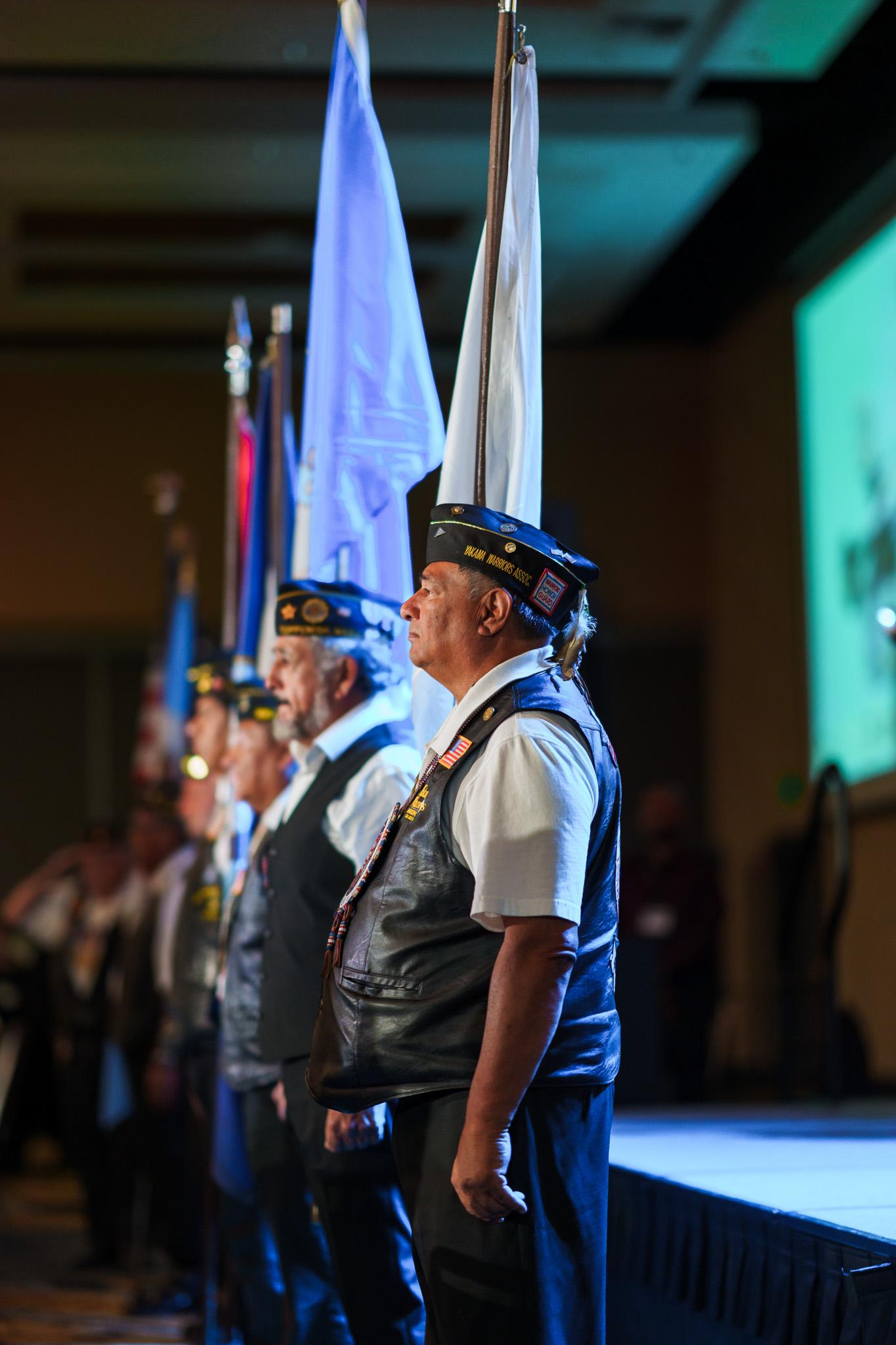 A line of indigenous veterans stand holding various flags as if they were leading a salute.