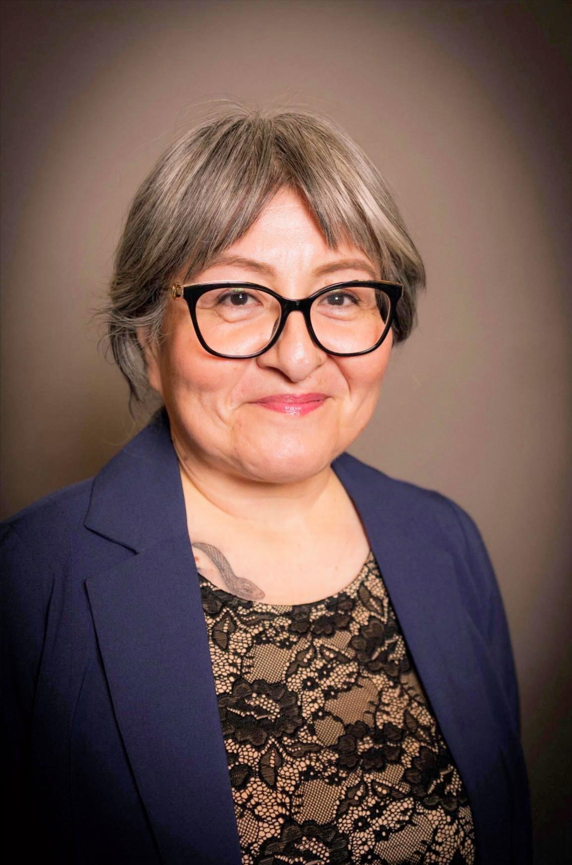 Yeni, a woman, is pictured in front of a tan wall wearing a blue jacket over a brown shirt. Her short grey hair is worn loose about her face and she is smiling.  