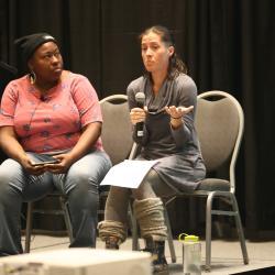 Two people sit on chairs on a platform speaking. One of them holds a microphone and notes.