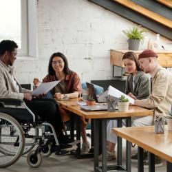 Two pairs of employees working in small groups by workplace while female economist pointing at document held by male colleague in wheelchair